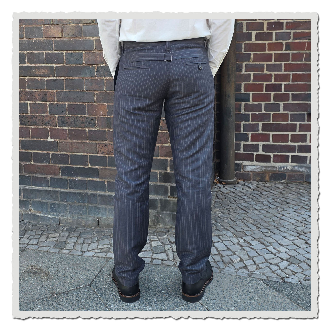 Rückansicht einer grauen Nadelstreifenhose am Model vor Backsteinwand und Gehweg im Außenbereich.